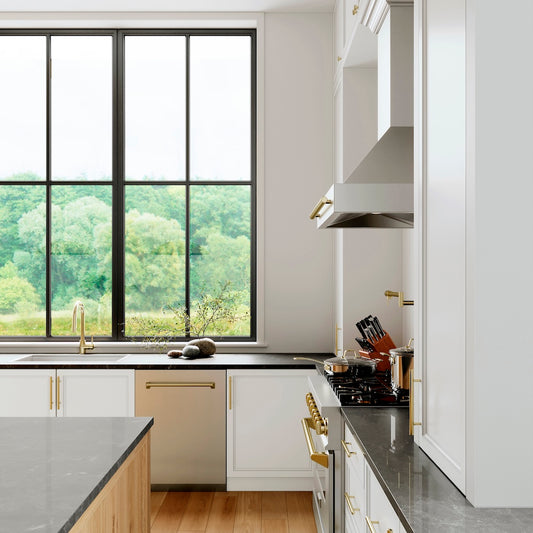 ZLINE Autograph Edition 24 in. Tallac Series 3rd Rack Top Control Built-In Tall Tub Dishwasher in Stainless Steel with Polished Gold Handle, 51dBa (DWVZ-304-24-G) in a kitchen with white cabinets, black countertops, and a large window view of greenery.