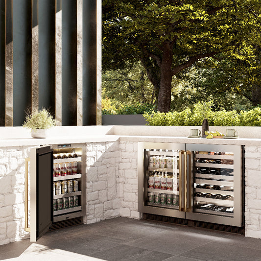 ZLINE Autograph Edition 24 in. Touchstone 151 Can Beverage Fridge With Stainless Steel Glass Door And Polished Gold Handle (RBSOZ-GS-24-G) in an outdoor kitchen with a matching built-in wine cooler against a stone wall and greenery.