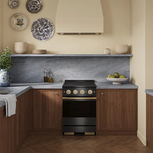 A modern kitchen scene featuring a ZLINE Autograph Edition 24 in. 3.2 cu. ft. Marino Induction Range with 3 Element Cooktop and Electric Convection Oven in Black Stainless Steel with Champagne Bronze Accents (MINRBZ-24-CB) set into warm wood cabinets with a gray stone backsplash and countertop.