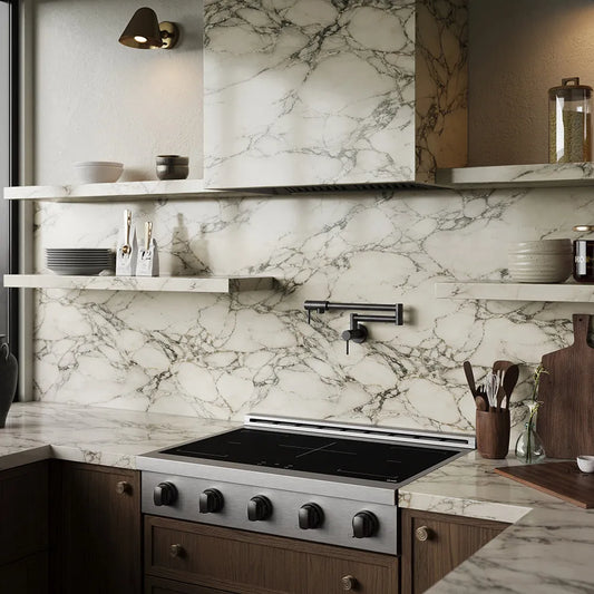 A modern kitchen interior featuring a white marble backsplash with dramatic gray veining, matching marble countertops, dark wood cabinetry, a ZLINE Autograph Edition 36 in. Paramount Induction Rangetop with 5 Element Cooktop in Stainless Steel and Matte Black Accents (SINRTZ-36-MB), floating open shelves with ceramic dishes, and a matte black faucet.