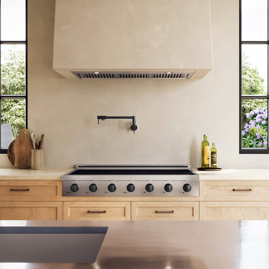 A ZLINE Autograph Edition 48 in. Paramount Induction Rangetop with 7 Element Cooktop in Stainless Steel and Matte Black Accents (SINRTZ-48-MB) set into a light wood kitchen island beneath a large cream-colored range hood. The kitchen features a textured beige backsplash, a black faucet, and windows showing green foliage outside.