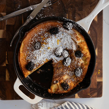 Skillet with a Dutch baby topped with berries and powdered sugar on a wooden cutting board.