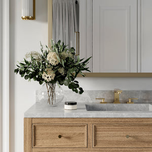 ZLINE Crystal Bay Widespread Bath Faucet in Polished Gold (CBY-BF-PG) on a bathroom vanity with wooden cabinets, marble countertop, and decorative flowers.