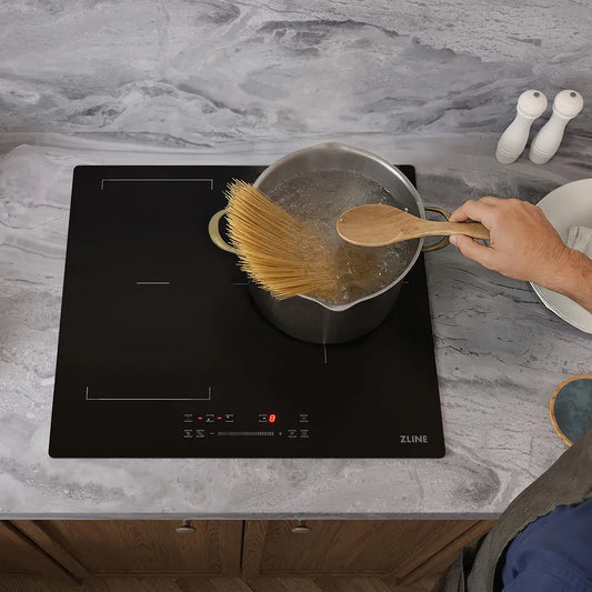 A ZLINE 24 in. Induction Cooktop with Anti-Scratch Glass and 3 Elements (FCIND-24) on a gray marble countertop, with a person's hand holding a wooden spoon over a pot of boiling water. A pasta brush is also resting on the cooktop.