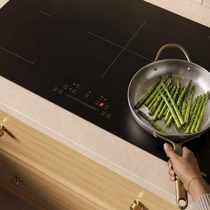 A stainless steel frying pan with asparagus cooking inside sits on a ZLINE 36 in. Induction Cooktop with Anti-Scratch Glass and 5 Elements (FCIND-36), with a hand holding the pan handle. The stovetop is installed in a light wood kitchen cabinet with a cream countertop.