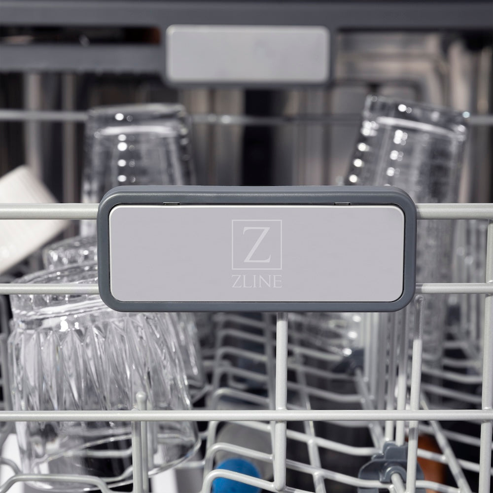 ZLINE Autograph Edition 24 in. Monument Series 3rd Rack Top Touch Control Tall Tub Dishwasher in Custom Panel Ready with Matte Black Handle, 45dBa (DWMTZ-24-MB) logo on dish rack with dishes in the background.