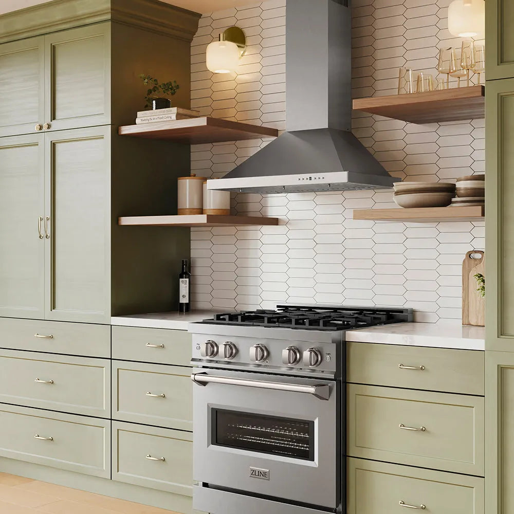 A modern kitchen scene featuring a stainless steel gas range and ZLINE 30 in. Convertible Vent Wall Mount Range Hood in Stainless Steel (KB-30), set against a white hexagonal tile backsplash and light sage green cabinets with open wood shelves.