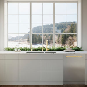 ZLINE Autograph Edition 24 in. Monument Series 3rd Rack Dishwasher with Auto Door Open Drying, 44dBa, Stainless Steel Panel with Polished Gold Handle (DWMTZ-304-24-G) in a kitchen with large window view of a lake and trees