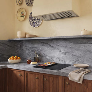 A kitchen scene with a gray marble-look backsplash and matching countertop, warm wood cabinets, a stainless range hood, decorative wall plates, and food cooking on a ZLINE 24 in. Induction Cooktop with Anti-Scratch Glass and 3 Elements (FCIND-24).