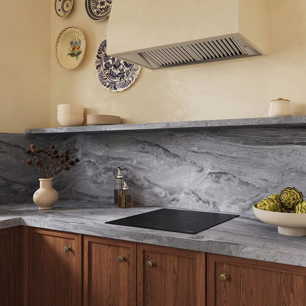 A kitchen interior with a gray marble-look countertop and matching marble backsplash, warm wood cabinets, a ZLINE 24 in. Induction Cooktop with Anti-Scratch Glass and 3 Elements (FCIND-24), cream walls, decorative plates, and a vase with dried stems.