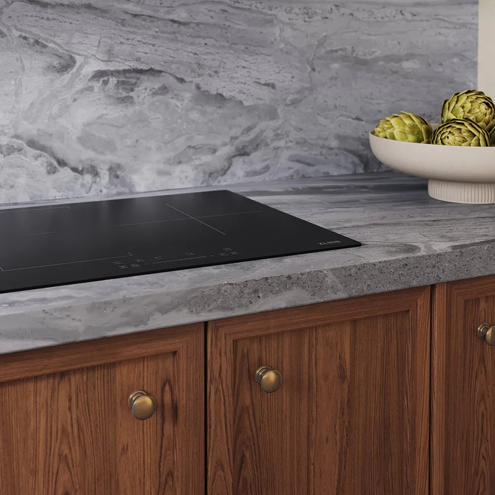 ZLINE 24 in. Induction Cooktop with Anti-Scratch Glass and 3 Elements (FCIND-24) embedded in a gray marble countertop, set against a matching gray marble backsplash and wooden cabinets with brass knobs. A white bowl of artichokes sits on the counter.