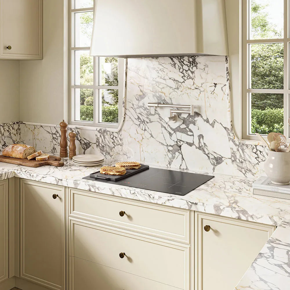 Cream-colored kitchen cabinets with a matching marble-look backsplash and countertop, featuring a ZLINE 30 in. Induction Cooktop with Anti-Scratch Glass and 4 Elements (FCIND-30), stainless steel range hood, and windows with white frames.