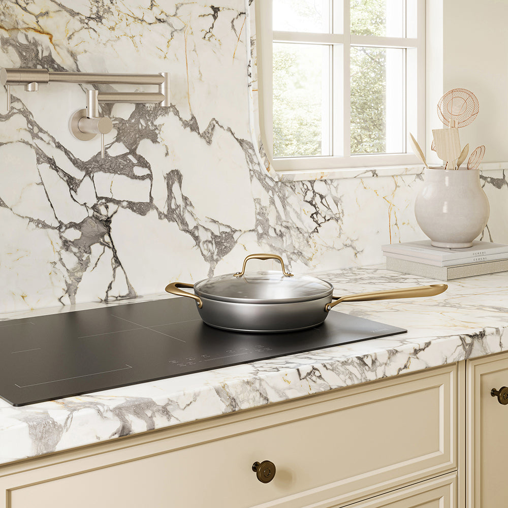 A ZLINE 30 in. Induction Cooktop with Anti-Scratch Glass and 4 Elements (FCIND-30) with a silver pan and lid on a white and gray marble countertop in a bright kitchen setting.