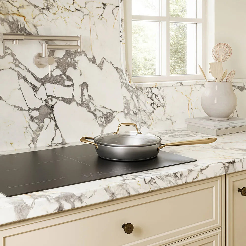 A ZLINE 30 in. Induction Cooktop with Anti-Scratch Glass and 4 Elements (FCIND-30) with a silver pan and lid on a white and gray marble countertop in a bright kitchen setting.