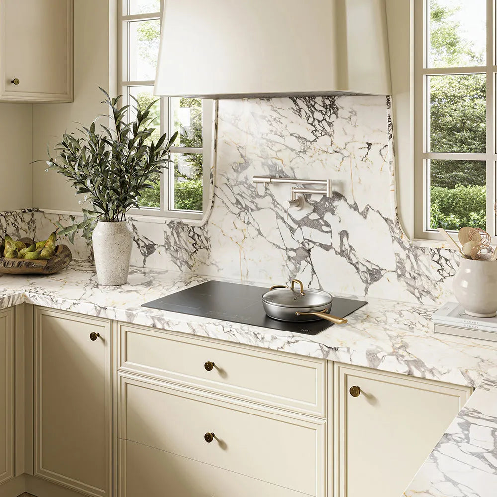 Cream-colored kitchen cabinetry with a matching marble-look backsplash and countertop, featuring a ZLINE 30 in. Induction Cooktop with Anti-Scratch Glass and 4 Elements (FCIND-30) and a silver pot, with a vase of greenery and windows in the background.