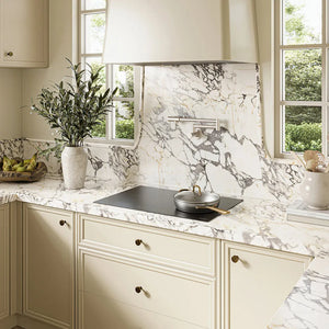 Cream-colored kitchen cabinetry with a matching marble-look backsplash and countertop, featuring a ZLINE 30 in. Induction Cooktop with Anti-Scratch Glass and 4 Elements (FCIND-30) and a silver pot, with a vase of greenery and windows in the background.