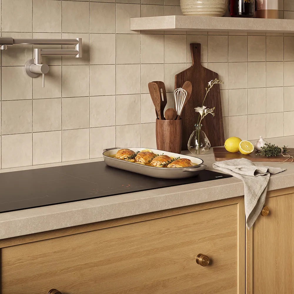 A kitchen scene with beige square wall tiles, a light wood countertop, a ZLINE 36 in. Induction Cooktop with Anti-Scratch Glass and 5 Elements (FCIND-36), wooden cabinets, and kitchen accessories including a baking dish with food, utensils, lemons, and a cloth.