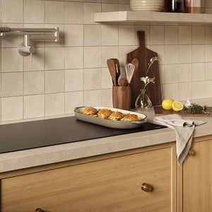 A kitchen scene with beige square wall tiles, a light wood countertop, a ZLINE 36 in. Induction Cooktop with Anti-Scratch Glass and 5 Elements (FCIND-36), wooden cabinets, and kitchen accessories including a baking dish with food, utensils, lemons, and a cloth.