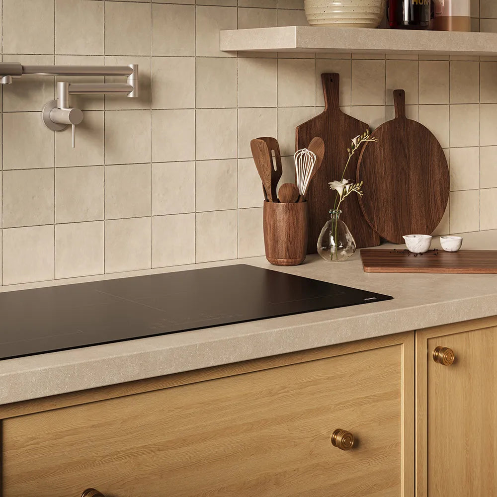 A kitchen scene with a ZLINE 36 in. Induction Cooktop with Anti-Scratch Glass and 5 Elements (FCIND-36), beige tiled backsplash, light wood cabinets, and dark wood cutting boards and utensils on the counter.