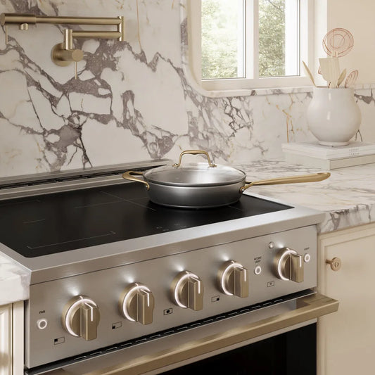 A ZLINE Autograph Edition 30 in. 4.2 cu. ft. Marino Induction Range with 4 Element Cooktop and Electric Convection Oven in Stainless Steel with Champagne Bronze Accents (MINRZ-30-CB) set in a kitchen with a marble backsplash and countertop.