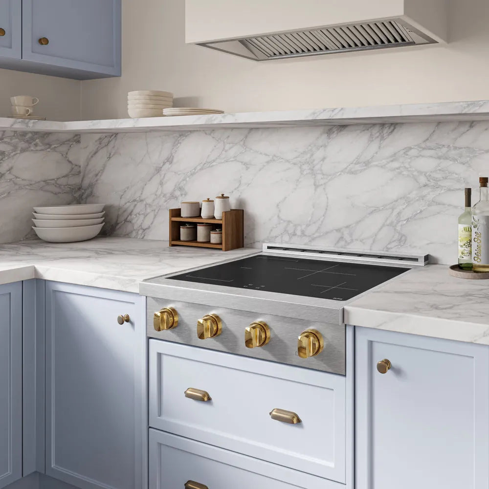 A luxury kitchen scene featuring a ZLINE Autograph Edition 30 in. Paramount Induction Rangetop with 4 Element Cooktop in Satin Stainless Steel and Polished Gold Accents (SINRTSZ-30-G) set into light blue cabinets with brass handles. The countertop is made of white marble with gray veining, and a matching marble backsplash is visible behind it.