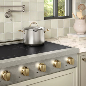 ZLINE Autograph Edition 36 in. Paramount Induction Rangetop with 5 Element Cooktop in Satin Stainless Steel and Champagne Bronze Accents (SINRTSZ-36-CB) in a designer kitchen with marble backsplash, light blue cabinets, angled.