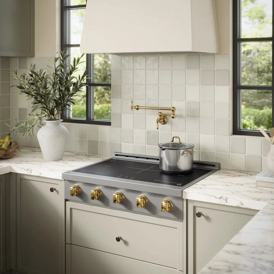 A custom kitchen scene featuring a ZLINE Autograph Edition 36 in. Paramount Induction Rangetop with 5 Element Cooktop in Stainless Steel and Polished Gold Accents (SINRTZ-36-G) set into sage green cabinets with matching marble countertops. A white subway tile backsplash and a brass wall-mounted faucet are visible behind the stove, with a white range hood and windows in the background.
