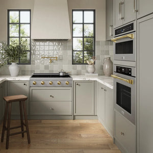 A modern kitchen interior with light sage green cabinets, a black gas range with gold knobs, ZLINE Autograph Edition 36 in. Paramount Induction Rangetop with 5 Element Cooktop in Stainless Steel and Polished Gold Accents (SINRTZ-36-G), a tiled backsplash, wood flooring, and a wooden stool.
