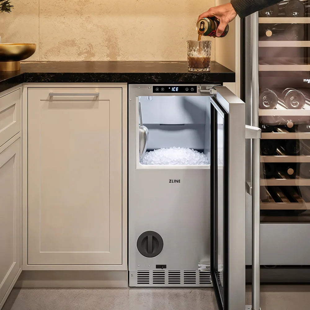 A built-in ZLINE 15 in. Touchstone Nugget Ice Maker with Drain Pump and Solid Stainless Steel Door (INGD-ST-15) in a modern kitchen setting, with a person pouring a dark beverage from a can into a glass on the counter.