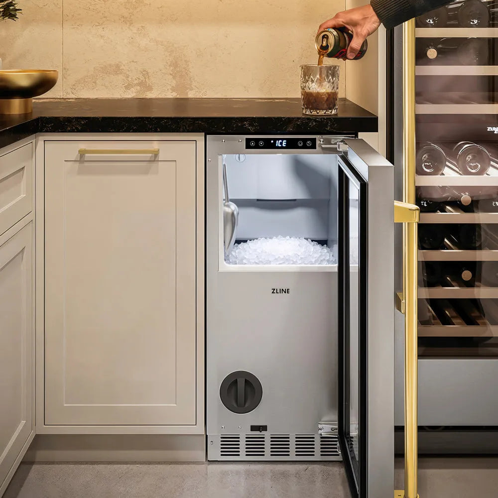 A ZLINE Autograph Edition 15 in. Touchstone Nugget Ice Maker with Drain Pump and Solid Stainless Steel Door with Polished Gold Handle (INGDZ-ST-15-G) shown with its door open as a hand pours a dark beverage from a can into a glass on the countertop.