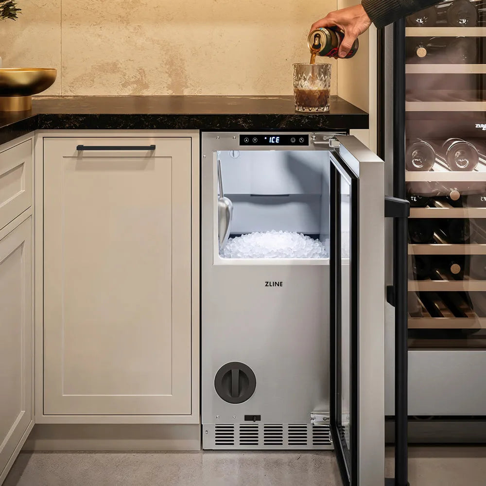A ZLINE Autograph Edition 15 in. Touchstone Nugget Ice Maker with Drain Pump and Solid Stainless Steel Door with Matte Black Handle (INGDZ-ST-15-MB), with its door open, showing a fresh ice tray. A hand is pouring a dark soda from a can into a glass on the black countertop next to the fridge.