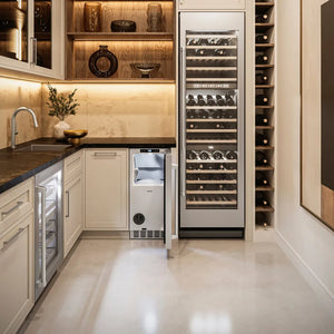 A modern wine refrigerator with a glass door stands in a luxury wine cellar setting next to a built-in wine rack and under-cabinet ZLINE 15 in. Touchstone Nugget Ice Maker with Drain Pump and Solid Stainless Steel Door (INGD-ST-15).
