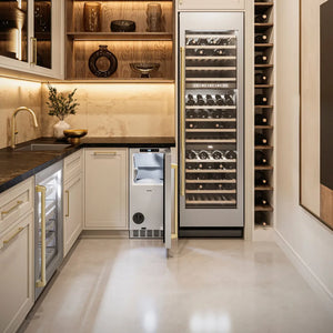 A luxury wine cellar featuring a stainless steel wine refrigerator with an integrated ZLINE Autograph Edition 15 in. Touchstone Nugget Ice Maker with Drain Pump and Solid Stainless Steel Door with Champagne Bronze Handle (INGDZ-ST-15-CB), a built-in wine cooler, and a tall wood shelving unit next to it.