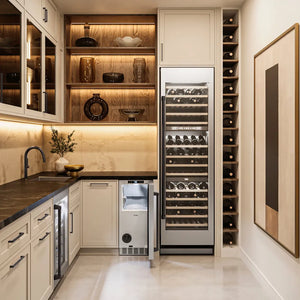 A modern wine cellar interior featuring a stainless steel wine refrigerator with glass doors, a vertical wall-mounted wine rack, ZLINE Autograph Edition 15 in. Touchstone Nugget Ice Maker with Drain Pump and Solid Stainless Steel Door with Matte Black Handle (INGDZ-ST-15-MB), dark countertops, warm wood shelving with under-cabinet lighting, and cream cabinetry.