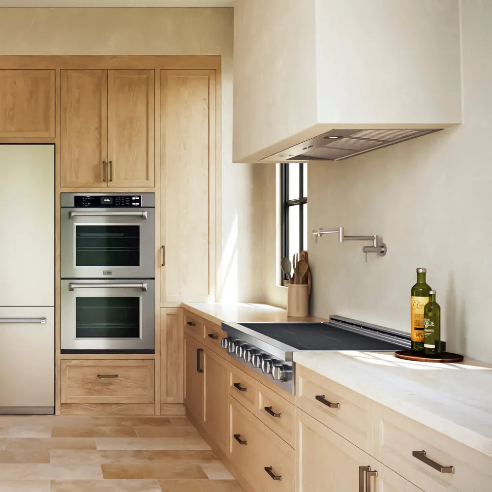 A ZLINE 48 in. Paramount Induction Rangetop with 7 Element Cooktop in Stainless Steel (SINRT48) in a designer kitchen interior with light wood cabinetry, stainless steel built-in ovens and range hood, white countertops, and a few kitchen items on the counter.