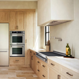 A ZLINE 48 in. Paramount Induction Rangetop with 7 Element Cooktop in Stainless Steel (SINRT48) in a designer kitchen interior with light wood cabinetry, stainless steel built-in ovens and range hood, white countertops, and a few kitchen items on the counter.