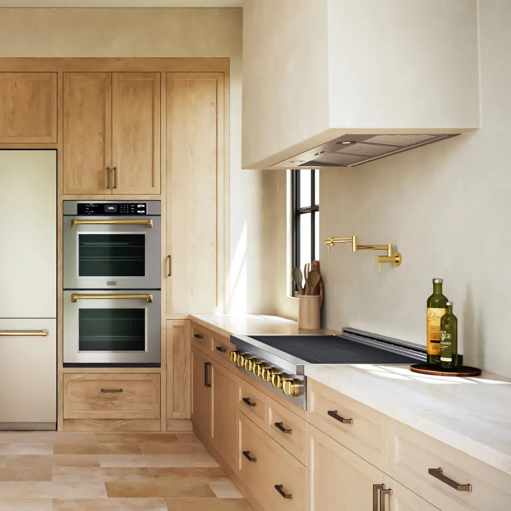 A modern kitchen interior with light wood cabinetry, stainless steel built-in ovens, a ZLINE Autograph Edition 48 in. Paramount Induction Rangetop with 7 Element Cooktop in Stainless Steel and Polished Gold Accents (SINRTZ-48-G), white countertops, and brass hardware. A bottle and a glass sit on the counter near a window.