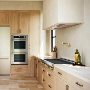 A modern kitchen interior with light wood cabinetry, stainless steel built-in ovens, a ZLINE Autograph Edition 48 in. Paramount Induction Rangetop with 7 Element Cooktop in Stainless Steel and Polished Gold Accents (SINRTZ-48-G), white countertops, and brass hardware. A bottle and a glass sit on the counter near a window.