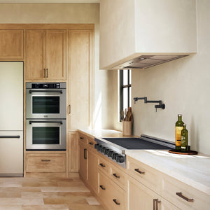 A ZLINE Autograph Edition 48 in. Paramount Induction Rangetop with 7 Element Cooktop in Stainless Steel and Matte Black Accents (SINRTZ-48-MB) in a modern kitchen interior with light wood cabinetry, white countertops, a built-in double oven, and a range hood. A bottle of olive oil and a green bottle sit on the counter near a window.