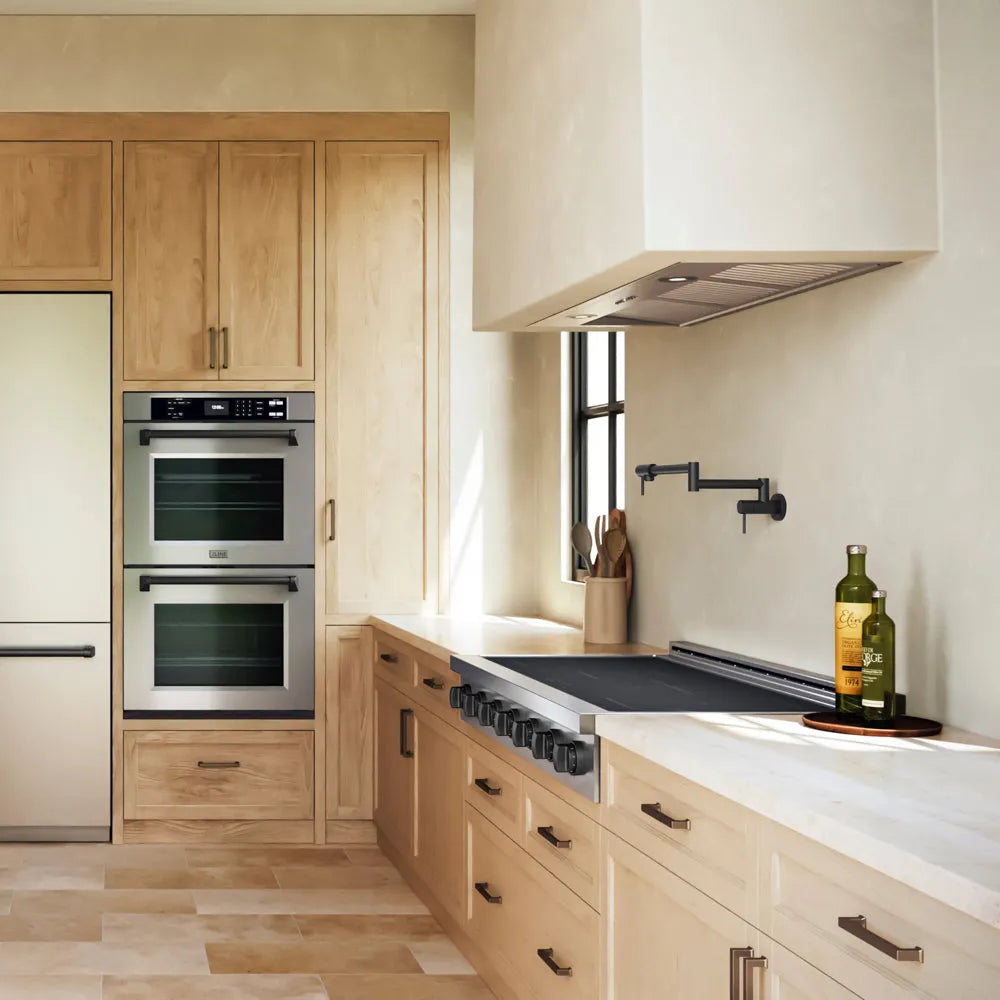 A ZLINE Autograph Edition 48 in. Paramount Induction Rangetop with 7 Element Cooktop in Stainless Steel and Matte Black Accents (SINRTZ-48-MB) in a modern kitchen interior with light wood cabinetry, white countertops, a built-in double oven, and a range hood. A bottle of olive oil and a green bottle sit on the counter near a window.