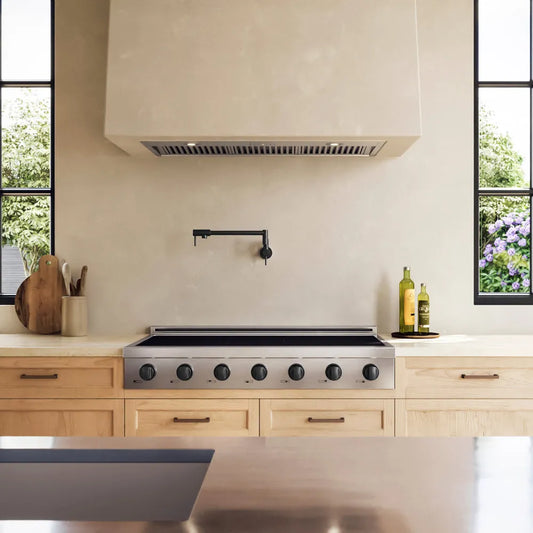 A ZLINE Autograph Edition 48 in. Paramount Induction Rangetop with 7 Element Cooktop in Stainless Steel and Matte Black Accents (SINRTZ-48-MB) set into a light wood kitchen island beneath a large cream-colored range hood. The kitchen features a textured beige backsplash, a black faucet, and windows showing green foliage outside.
