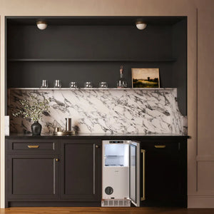 A ZLINE Autograph Edition 15 in. Touchstone Nugget Ice Maker with Drain Pump and Panel-Ready Door with Champagne Bronze Handle (INGDPZ-15-CB) with an open door, set into a dark charcoal kitchen cabinet beneath a white and gray marble backsplash.