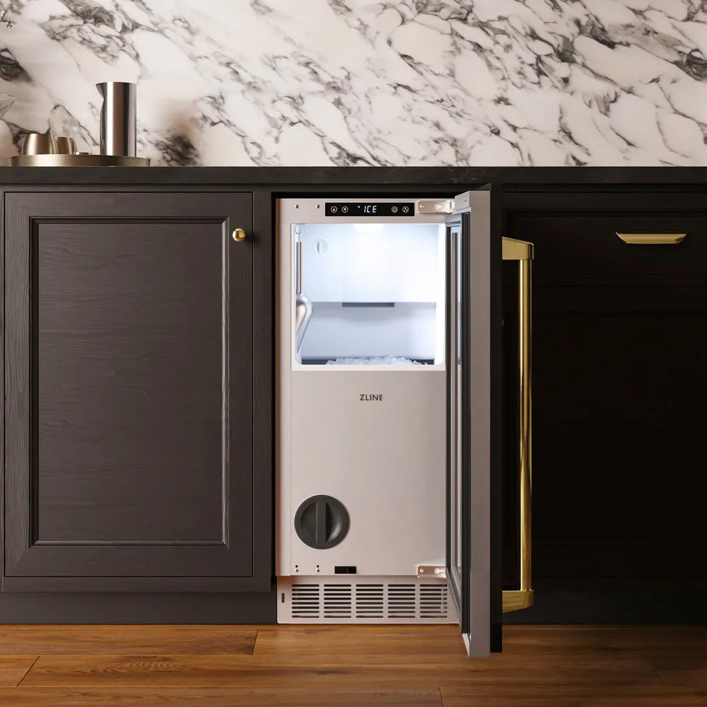 ZLINE Autograph Edition 15 in. Touchstone Nugget Ice Maker with Drain Pump and Panel-Ready Door with Polished Gold Handle (INGDPZ-15-G) with its door open, set into a kitchen cabinet with a dark countertop and marble backsplash.