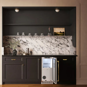 A ZLINE Autograph Edition 15 in. Touchstone Nugget Ice Maker with Drain Pump and Panel-Ready Door with Polished Gold Handle (INGDPZ-15-G)integrated into dark charcoal cabinetry with a white and gray marble backsplash. The scene is styled as a high-end bar or kitchen counter with glassware, a bottle, and decorative items.