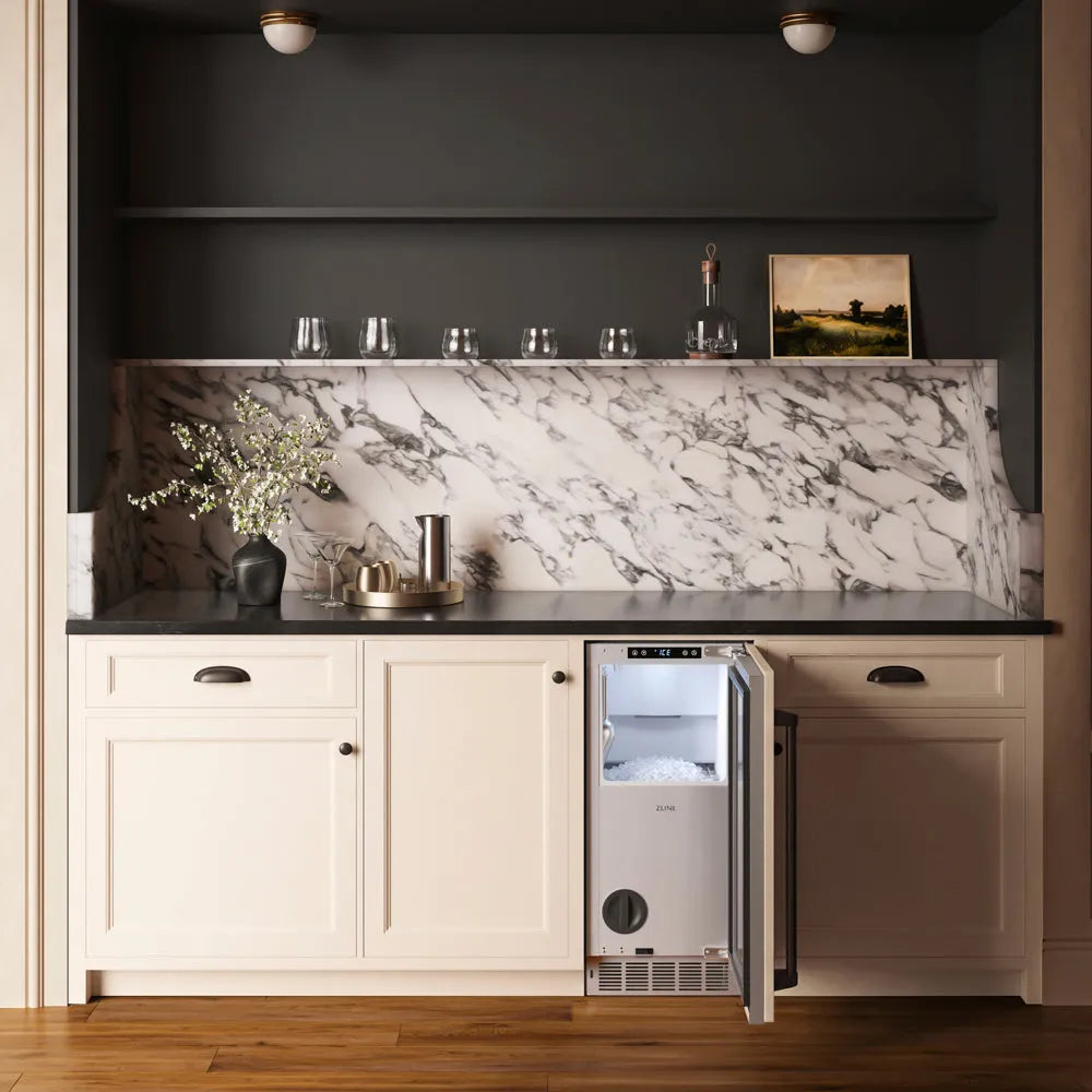 A built-in under-counter ZLINE Autograph Edition 15 in. Touchstone Nugget Ice Maker with Drain Pump and Panel-Ready Door with Matte Black Handle (INGDPZ-15-MB) with its door open, set into a cream-colored kitchen cabinet with a black countertop and a white marble backsplash.