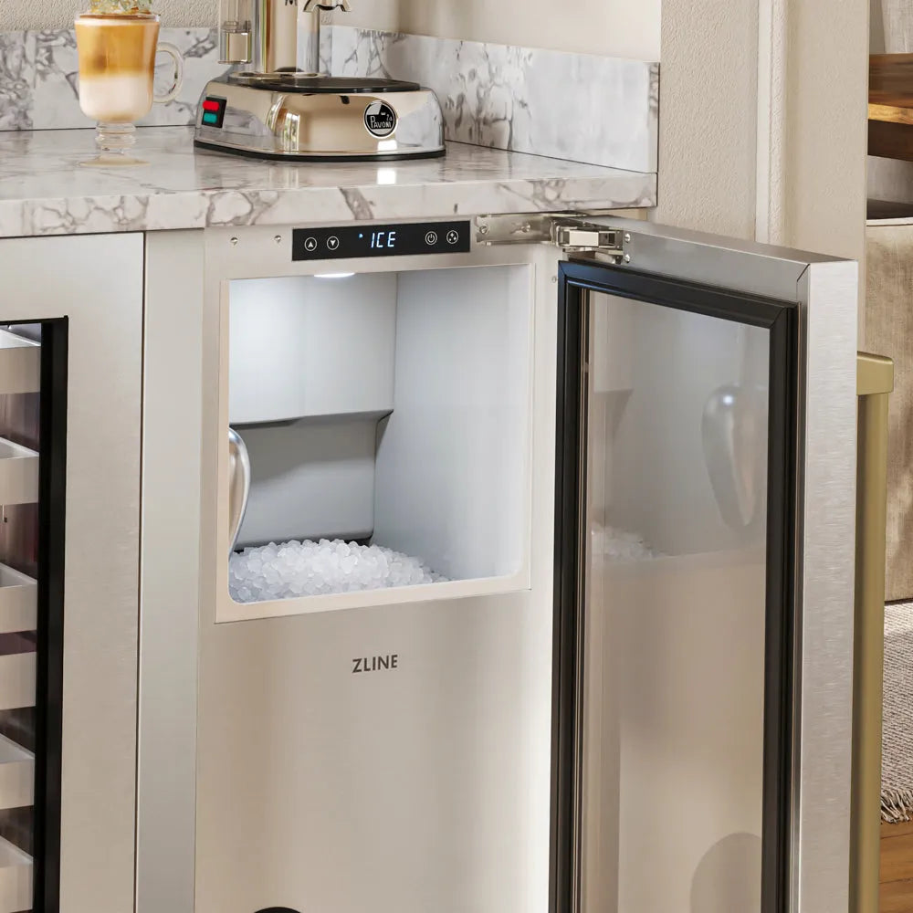 ZLINE Autograph Edition 15 in. Touchstone Nugget Ice Maker with Drain Pump and Solid Stainless Steel Door with Champagne Bronze Handle (INGDZ-ST-15-CB) with its door open, showing a digital display and a compartment filled with ice, installed under a marble countertop in a kitchen setting.