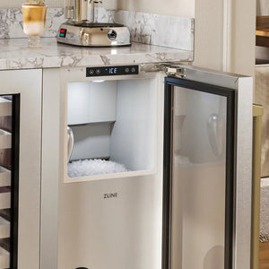 ZLINE Autograph Edition 15 in. Touchstone Nugget Ice Maker with Drain Pump and Solid Stainless Steel Door with Champagne Bronze Handle (INGDZ-ST-15-CB) with its door open, showing a digital display and a compartment filled with ice, installed under a marble countertop in a kitchen setting.