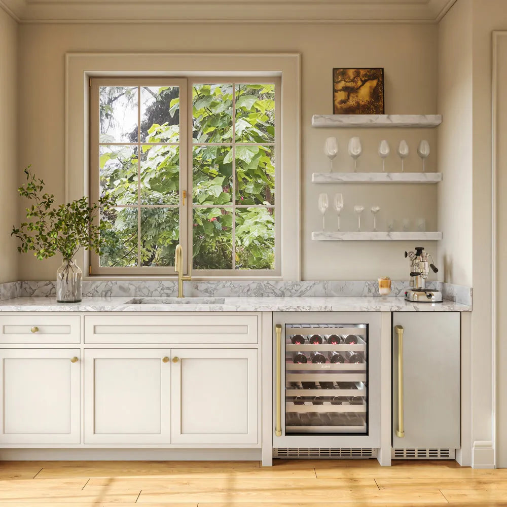 A modern kitchen cabinetry and appliance setup in a warm beige interior, featuring white cabinets, a marble countertop, a ZLINE Autograph Edition 15 in. Touchstone Nugget Ice Maker with Drain Pump and Solid Stainless Steel Door with Champagne Bronze Handle (INGDZ-ST-15-CB), and a window looking out onto green foliage.