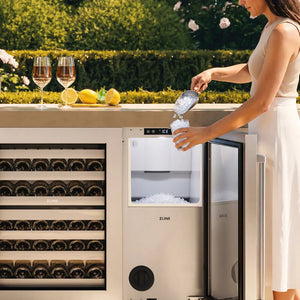 A woman in a white dress serves ice from a ZLINE 15 in. Touchstone Nugget Ice Maker with Drain Pump and Solid Stainless Steel Door (INGD-ST-15), set against a lush green garden background.