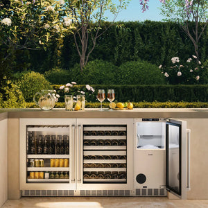 An outdoor kitchen with an open ZLINE 15 in. Touchstone Nugget Ice Maker with Drain Pump and Solid Stainless Steel Door (INGD-ST-15), set into a beige patio counter with a landscaped garden background.